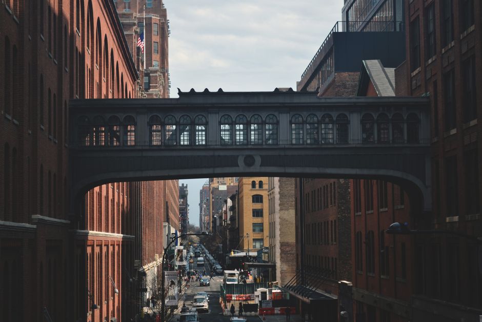 A street scene in an urban area featuring tall brick buildings on both sides. A decorative, enclosed pedestrian bridge with arched windows connects the buildings. The street below is filled with traff