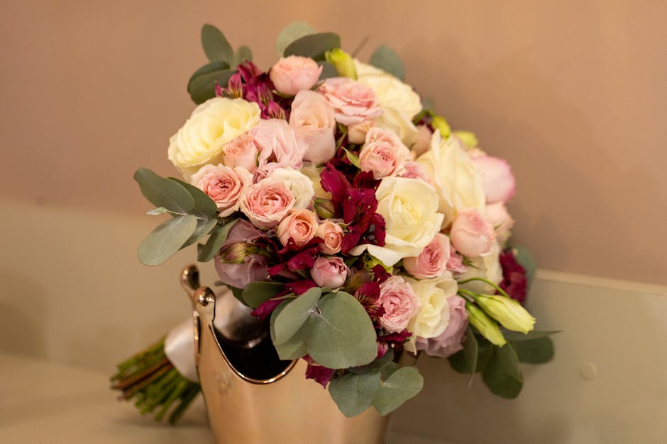 A close-up view of a fresh floral bouquet featuring pale pink spray roses, large white roses, and deep burgundy blooms, accented with lush green eucalyptus leaves. The bouquet is arranged in a rounded