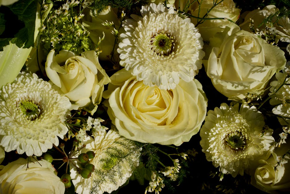 A close-up view of a floral bouquet featuring large, creamy white roses and white dahlias with layered petals. The arrangement includes delicate green foliage and small berry-like elements, all set ag