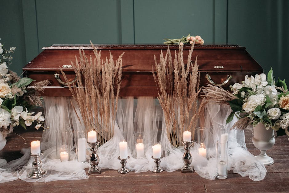 A funeral floral arrangement featuring two large white vases filled with white roses, orchids, and greenery, placed on either side of a polished wooden casket. In front of the casket, there are tall,