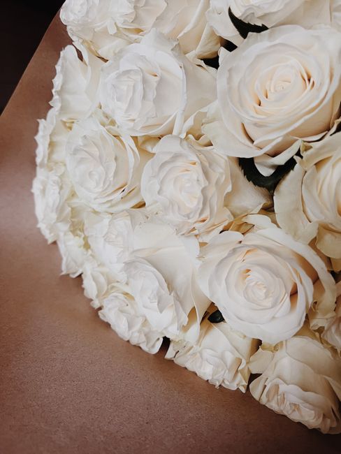 Close-up of a floral arrangement featuring creamy white roses and peonies with delicate, layered petals, arranged tightly together on a light brown surface. The flowers appear fresh, with lush petals