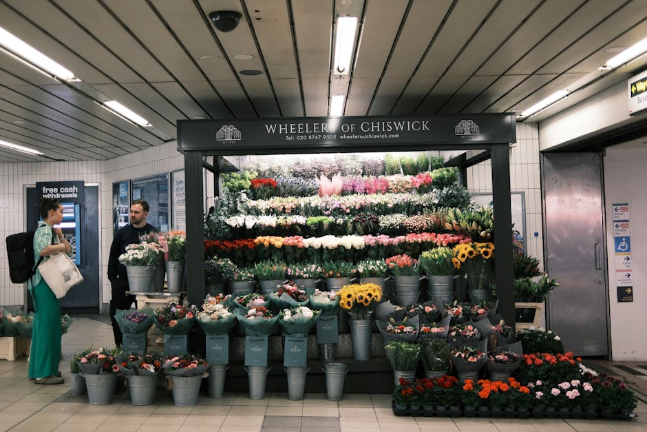 A vibrant flower stall at Fulham Broadway station operated by Wheeler of Chiswick, displaying an array of fresh flowers arranged in tiered layers. The arrangement includes roses, tulips, sunflowers, a